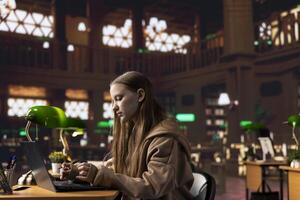 College student working on her laptop under a green lamp in a quiet setting, using the university library in the campus to prepare for an important school project. Young girl gathers data on her pc. photo