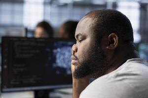 Tired man falling asleep while using artificial intelligence technology to optimize data center. Exhausted server hub expert taking break from using AI tech to streamline operations, slacking off photo