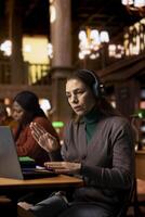 Mature woman attending a webinar from a quiet school library, taking notes during a digital lesson and engaging with her professor and classmates through wireless telecommunication tools. photo