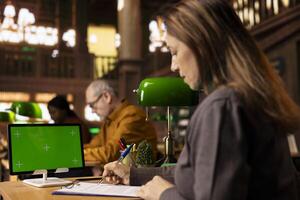 Wise adult learner focused taking notes for research next to chroma key, gathering information and sources for citations. Mature woman working on her diploma paperwork in a public library. photo