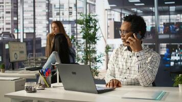 Businessman engages in a mobile phone conversation at his desk, managing a project and discussing deal making strategies. Multitasking skills drive productivity in a corporate setting. Camera B. photo