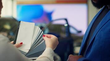 Saleswoman in dealership talking with client, offering color swatch samples choice, trying to grasp their preferences for vehicle. Agent providing customer with color wheel tinting selection, camera A photo