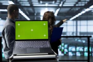 Isolated screen mockup laptop in front of technicians in blurry background supervising server room gear. Close up of chroma key notebook next to admins overseeing data center rigs photo