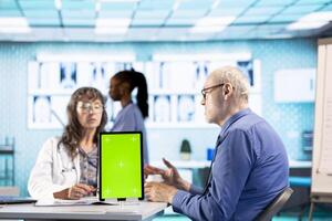Woman general practitioner uses device with green screen during check up visit with senior man, consulting patient on insurance at the facility. Old medic and client discuss wellness and recovery. photo