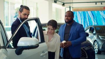 Car sales representative inviting clients to touch vehicle door and test quality, helping individuals make investment decision. Showroom agent asking customers to check automobile features, camera A photo