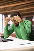 Man feeling clueless while preparing for school exam at home office desk, struggling to memorize info. African american student researching for college tests using laptop, doing brainstorming photo