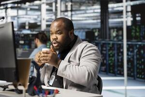 Data center technician drinking coffee analyzing software logs metrics on PC, optimizing storage systems. Server room employee enjoying beverage inspecting resource utilization on computer photo