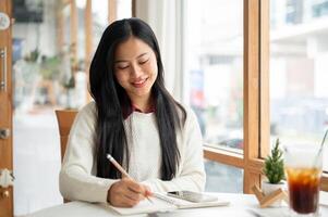 An asian woman is smiling while writing in notebook, sitting with a tablet and a drink on the table. photo
