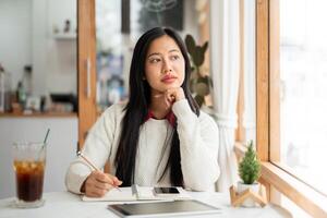 An asian woman is thinking while writing with pencil in notebook, with phone and tablet on the table photo
