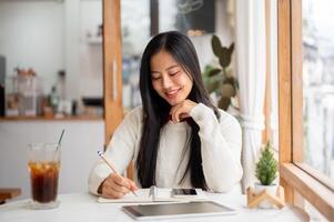 An asian woman is smiling while writing with a pencil on notebook with phone and tablet on the table photo