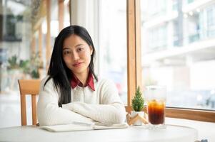 An asian woman is sitting with arm crossed over an open notebook while looking straight into camera. photo