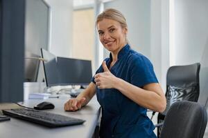 Nurse on Duty working on computer at the Reception Desk in modern clinic and showing thumb up photo