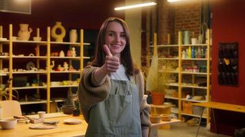 Caucasian woman girl smiling portrait in craft store glad happy female sculptor small business owner potter artist designer in art studio looking at camera thumb up gesture pottery workshop handmade video