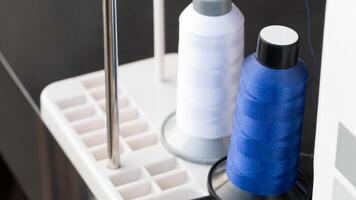 Two spools of embroidery thread. One bright blue and one white sitting on a white thread stand. Blue spool appears to be actively feeding into an embroidery machine suggesting a sewing or crafting act photo