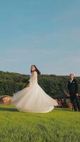 Bride Twirling in a White Gown Outdoors with Groom Watching. A bride twirls joyfully in her flowing white gown on a grassy field as the groom stands watching video