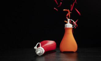 Bottles of chili sauce and ketchup are placed on the table with fresh red chili peppers, spicy food condiments, on a black and dark background. photo