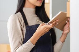 Close-up of young woman in apron taking notes in a notebook while managing inventory. photo