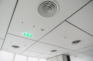 Clean modern office ceiling featuring air vents, recessed lighting, and grid pattern with emergency exit sign glowing green, creating bright and organized atmosphere photo