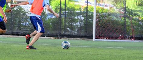 Soccer players in action on green grass field focusing on their legs and ball near goalpost during outdoor game with net and trees in background photo