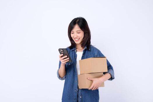 Southeast asian woman shopping online happily holds two cardboard boxes while checking her smartphone for orders or delivery updates in bright studio setting photo