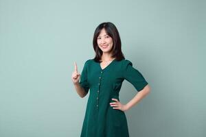 Southeast asian woman in green dress is smiling confidently while advertising with raised finger against plain light green background photo