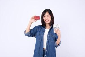 Southeast asian woman happily holds credit card and cash showing internet mobile application payment options with bright smile and casual denim shirt on white background photo