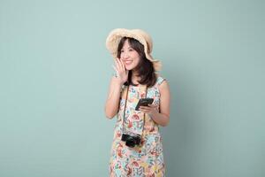 Southeast asian woman wearing floral dress and straw hat happily uses mobile phone with camera hanging around her neck while enjoying internet application on pastel background photo