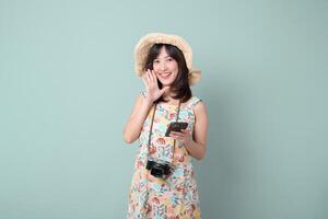 Southeast asian woman smiling happily while using mobile phone and internet application wearing floral dress and straw hat with camera hanging around her neck photo