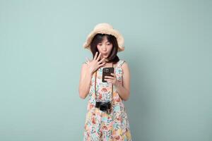 Southeast asian woman wearing floral dress and straw hat looks surprised while using mobile phone to browse internet application with camera hanging around her neck photo