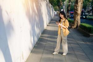 Southeast asian woman enjoys travel concept as she stands on sunny sidewalk using her phone with relaxed smile and casual outfit in urban outdoor setting photo