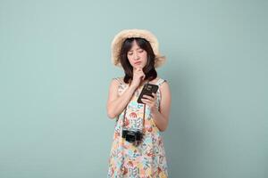 Thinking woman in summer dress and hat holding camera and mobile phone, standing on pastel green background, looking thoughtful photo