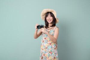 Southeast Asian woman wearing summer dress and straw hat smiles joyfully while holding camera against plain pastel background photo