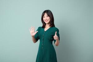 Southeast asian woman in green dress smiling and showing six fingers with one hand and thumbs up with other hand on plain background photo