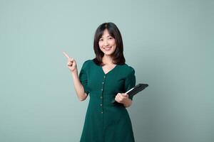 Southeast asian woman smiling while holding tablet and pointing upward, standing against plain background, representing internet technology and positive emotion photo