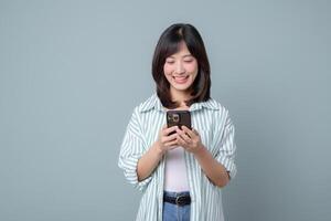 Southeast asian woman smiling while using smartphone in casual striped shirt standing against plain background showing happiness and modern lifestyle photo