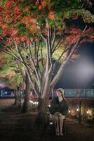 Southeast asian woman sits on bench under colorful autumn trees at night in peaceful park illuminated by soft lights creating calm and serene atmosphere photo