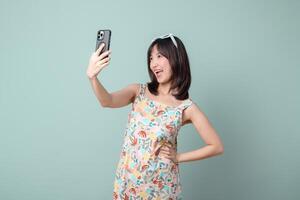 Southeast asian woman happily uses mobile phone to take selfie while enjoying internet application on plain background in colorful dress and sunglasses on her head photo