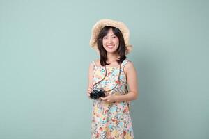 asian woman wearing floral dress and straw hat holds camera smiling happily, capturing travel concept with joy and excitement in simple studio setting photo