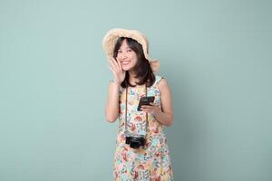 Southeast asian woman wearing hat smiles happily while holding mobile phone and camera, enjoying using internet application for travel and photography photo