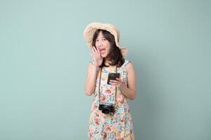 Southeast asian woman happily using mobile phone with internet application while wearing summer hat and floral dress with camera around her neck in studio photo