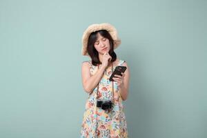 Southeast asian woman wearing hat looks thoughtfully at her mobile phone while holding camera around her neck using internet application for travel or photography photo