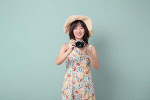 Southeast asian woman wearing floral dress and straw hat happily holding camera capturing travel moments in simple studio concept with soft green background photo