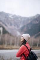 Southeast asian woman wearing warm hat and red jacket enjoys travel concept in mountains with black backpack, smiling peacefully in cold nature scene photo