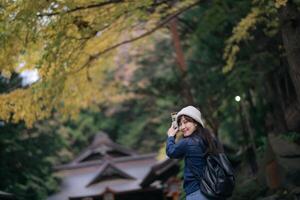 Southeast asian woman wearing white beanie and blue jacket smiles happily while holding phone in forested area with autumn leaves and traditional buildings in background photo