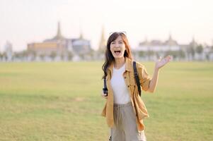 Southeast asian woman travel concept with joyful expression stands in open field waving happily with blurred cityscape in background during bright daylight photo