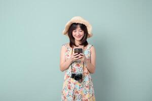 Southeast asian woman wearing floral dress and straw hat happily uses mobile phone to access internet application while camera hangs around her neck in front of plain background photo