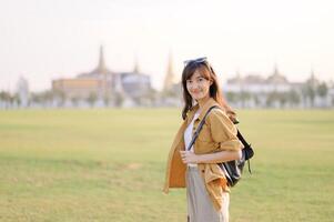 Southeast asian woman travel concept with casual outfit and backpack standing on green field with blurred temple background smiling happily photo