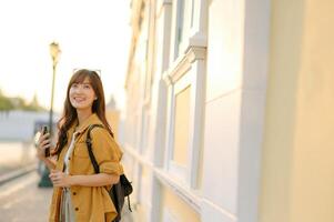 Southeast asian woman enjoys travel concept with bright smile holding phone outdoors near light colored building in warm sunlight creating happy and relaxed mood photo