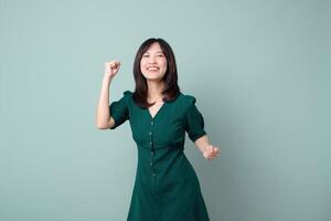 Southeast asian woman in green dress expressing joy and confidence for advertising with bright smile and raised fist on plain background photo