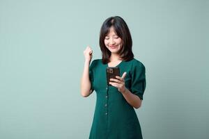 Southeast asian woman happily uses mobile internet application on her phone showing excitement and joy in simple green dress against plain background photo